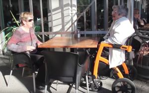 Dame in oranje rolstoel zit met haar dochter op een terras in de zon, waar blijft de koffie?